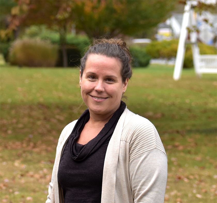 A woman in a dark shirt and cream cardigan against a fall background.